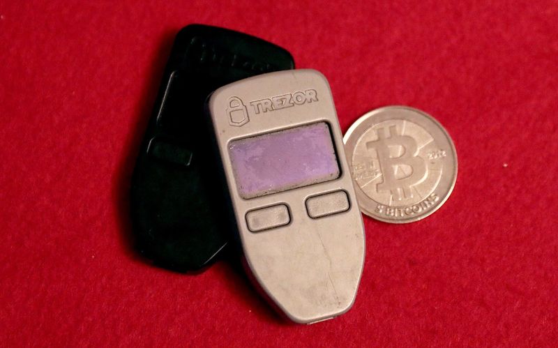 Dos billeteras físicas de criptomonedas de la marca Trezor junto a una moneda conmemorativa de bitcoin, sobre fondo rojo; no son equipos Passport