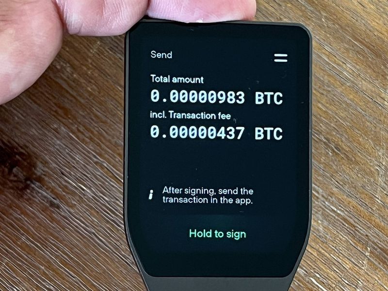 Mano que sostiene un monedero físico de criptomonedas de la marca Trezor, distinta de Keystone, con una transacción en pantalla